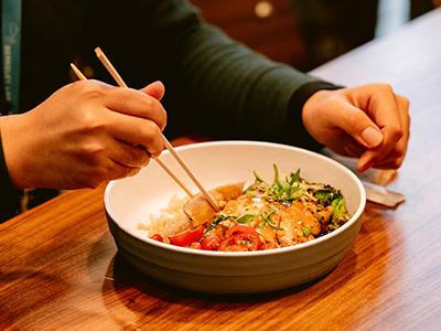Student eating bowl of chicken, rice and vegetables with chopsticks